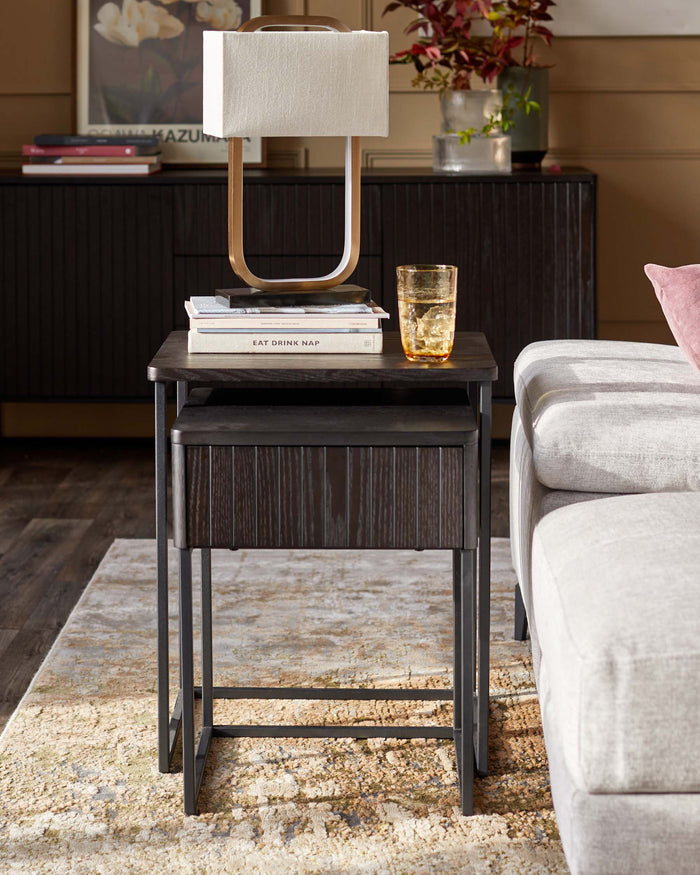 Dark wood nesting tables with a lamp and glass on top, next to a sofa, on a patterned rug.