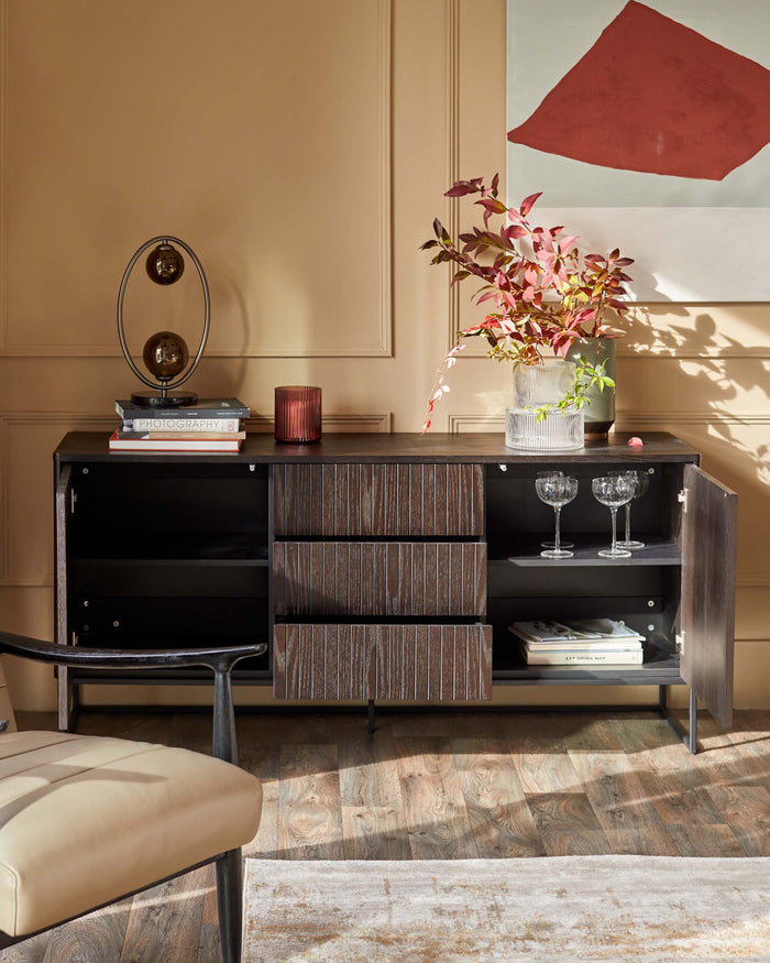 A dark wood sideboard with drawers, glassware, books and decor against a tan wall with modern artwork.