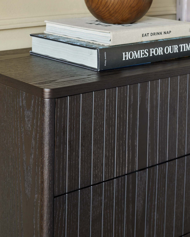 A dark wood cabinet with linear detailing, topped with books and a wooden bowl.