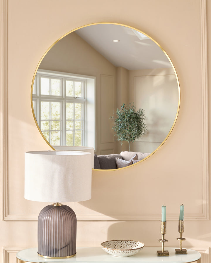 Large round gold-framed wall mirror reflecting a bright interior with windows, styled above a console table with lamp and decor.
