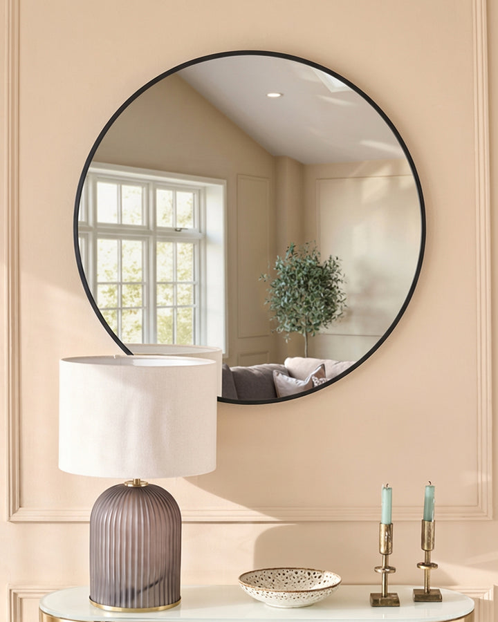 Large round black-framed wall mirror displayed above a white console table with decorative lamp and accessories.