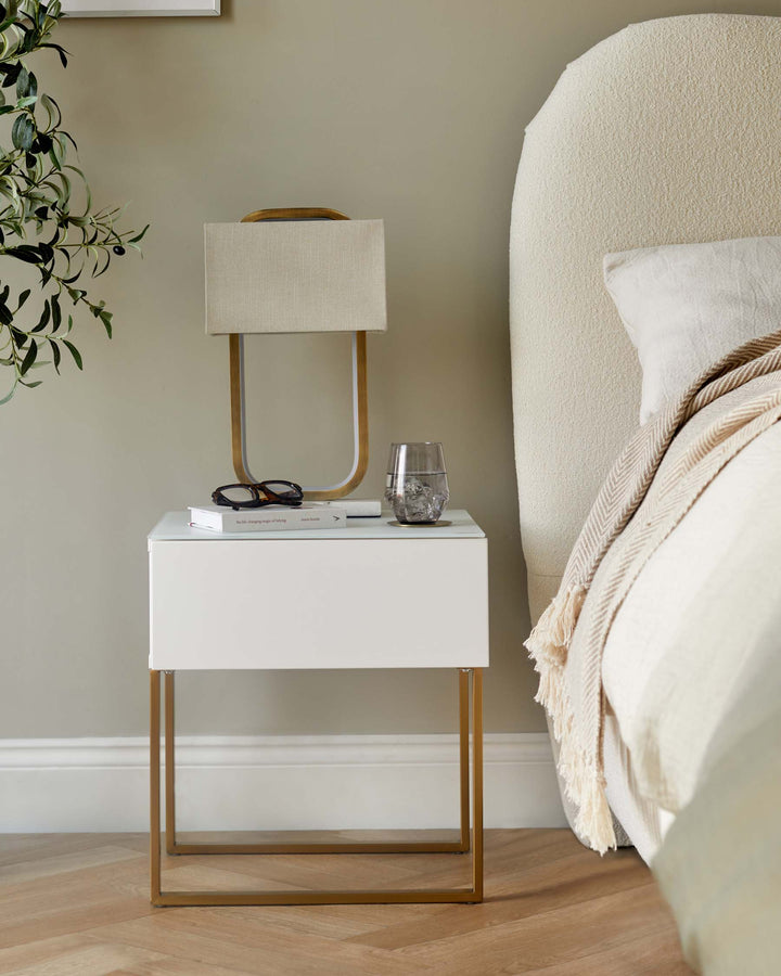 Bedside table with lamp, glass of water, and books, next to a bed with a neutral headboard and textured blanket.