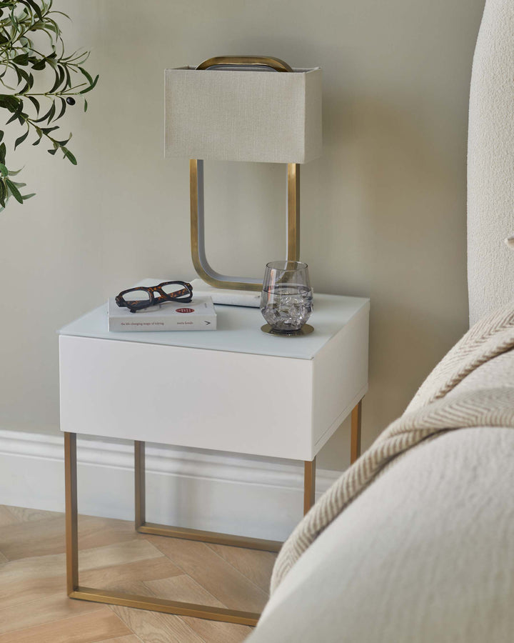 A white bedside table with gold frame. There is a lamp, book, glasses and a glass of water on top.