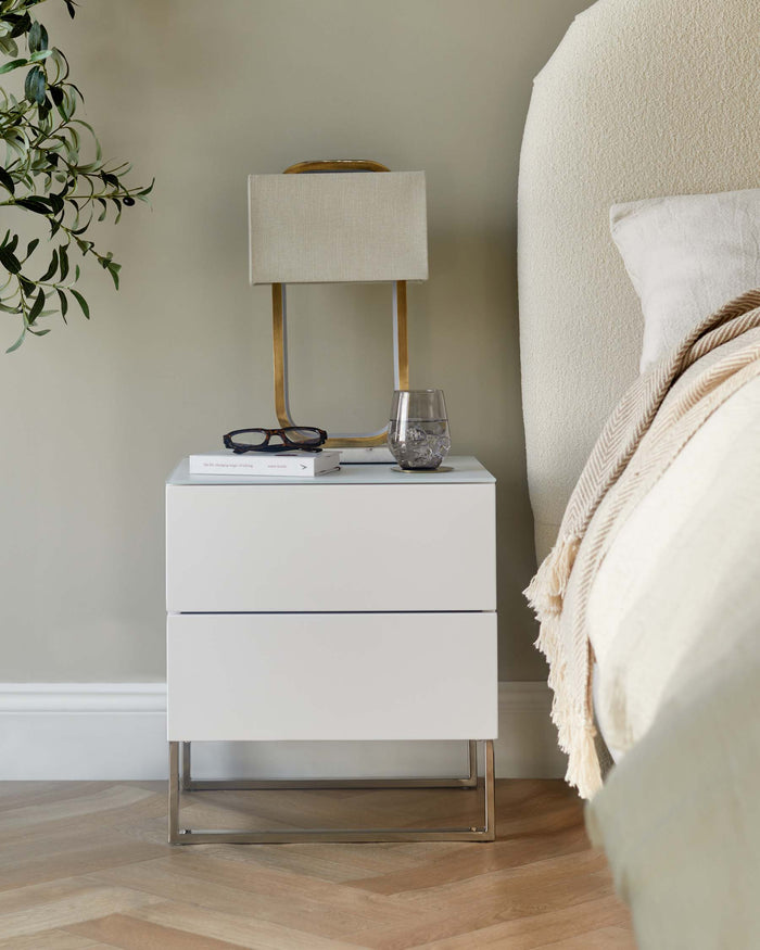A modern white bedside table with a metal frame, featuring drawers, a lamp, books and a glass of water.
