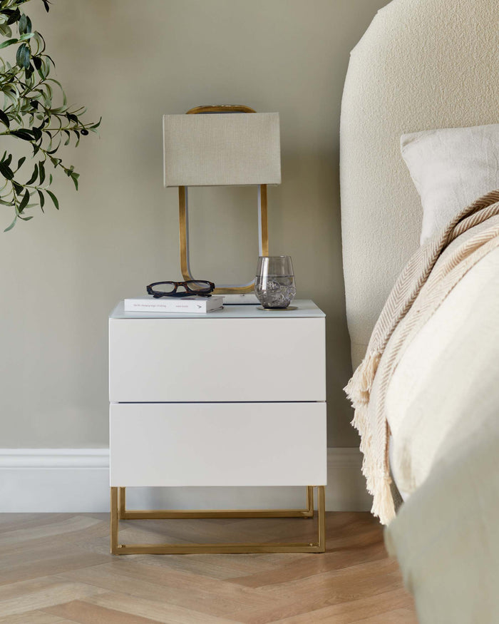 A white bedside table with gold legs, a neutral lamp, and bedhead in a bedroom setting.