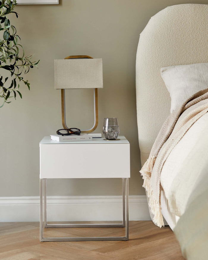 A stylish bedside table with a white drawer and chrome legs, complemented by a lamp, books, and a glass of water.