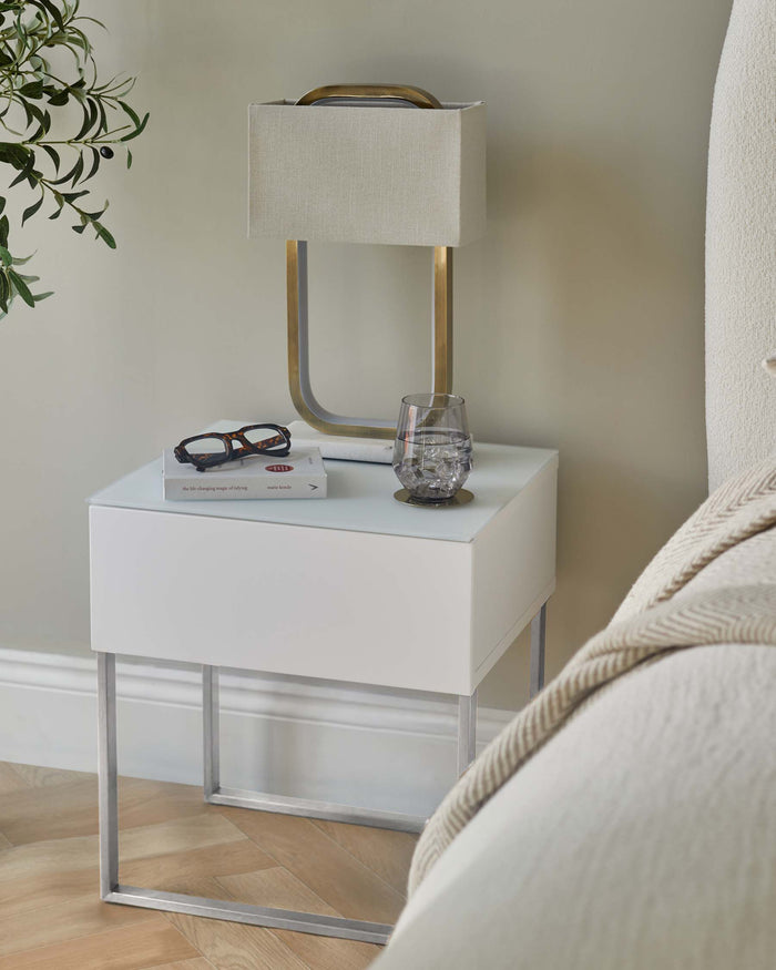 A modern bedside table with a white drawer, glass top and silver frame, complete with lamp, book, glasses and a glass of water.