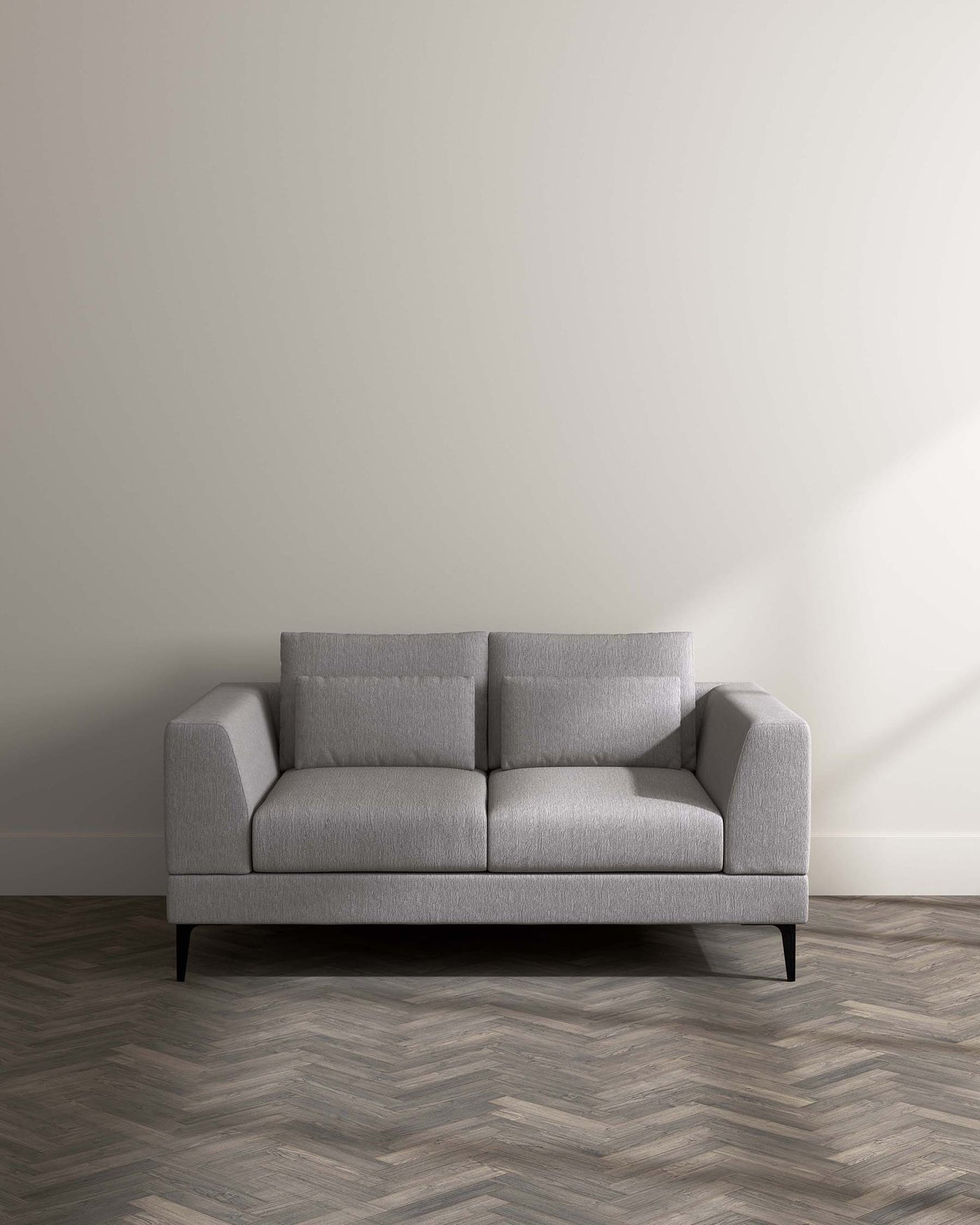 A modern three-seater sofa with light grey upholstery and minimalist design, featuring clean lines, square armrests, and slightly angled black metal legs. The sofa is placed on a herringbone pattern wood flooring against a plain light wall, showcasing a blend of contemporary style and comfort.