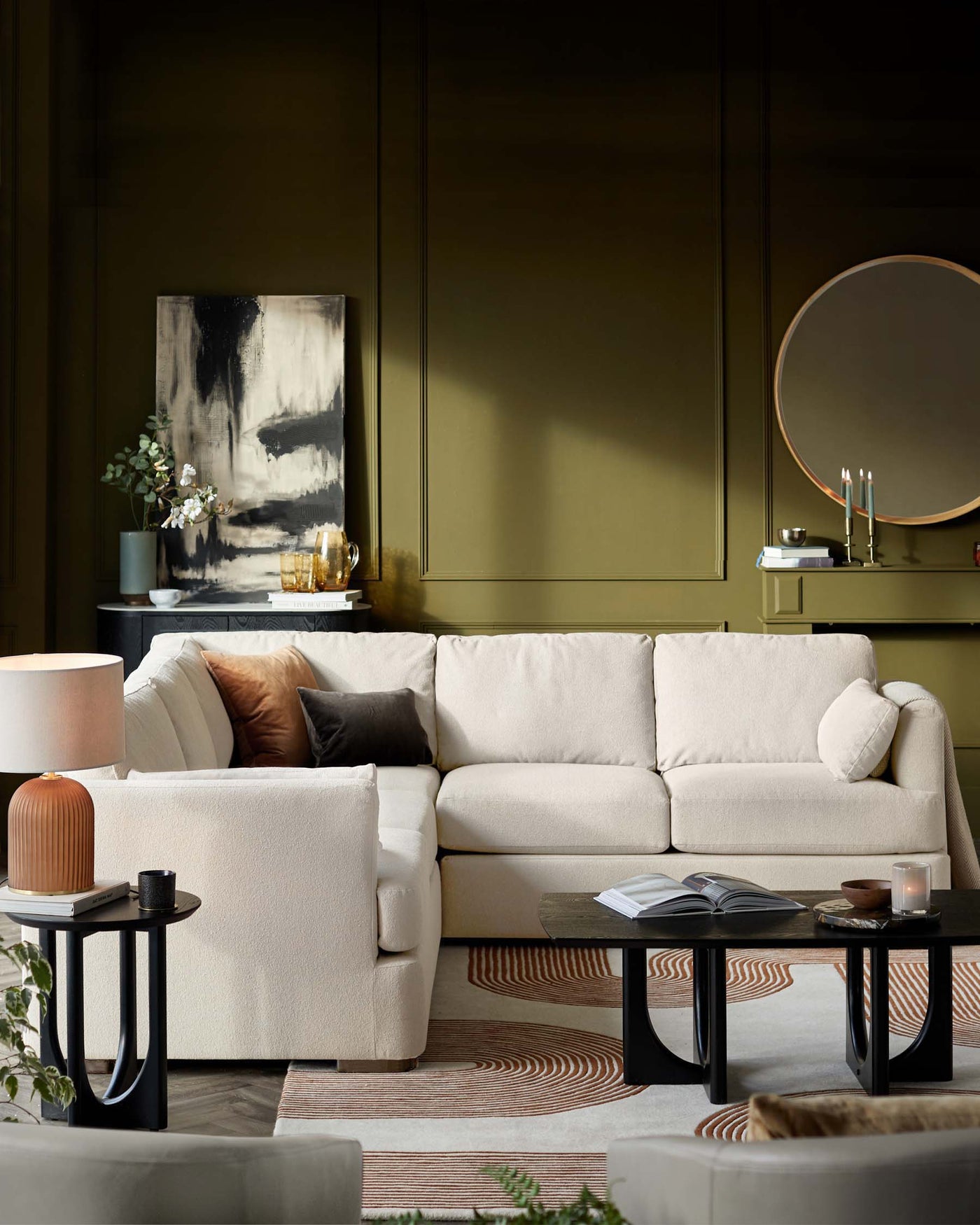 Modern L-shaped beige sectional sofa with plush cushions, paired with a round black side table and a rectangular black coffee table, set on a patterned rug in a sophisticated living room setting.