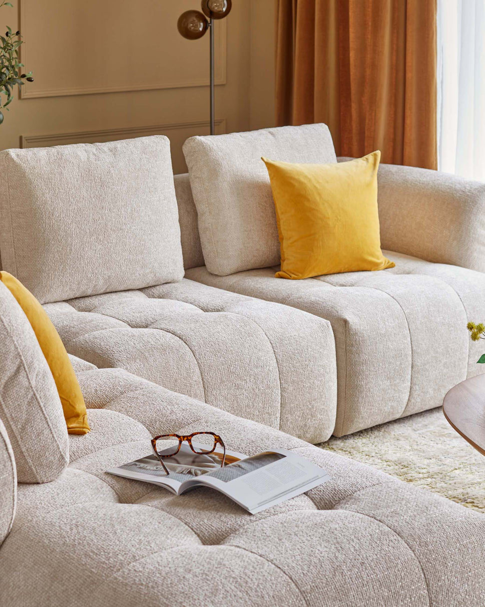 A stylish light beige sectional sofa with textured upholstery, featuring a bright yellow pillow and a coffee table with a magazine and glasses.