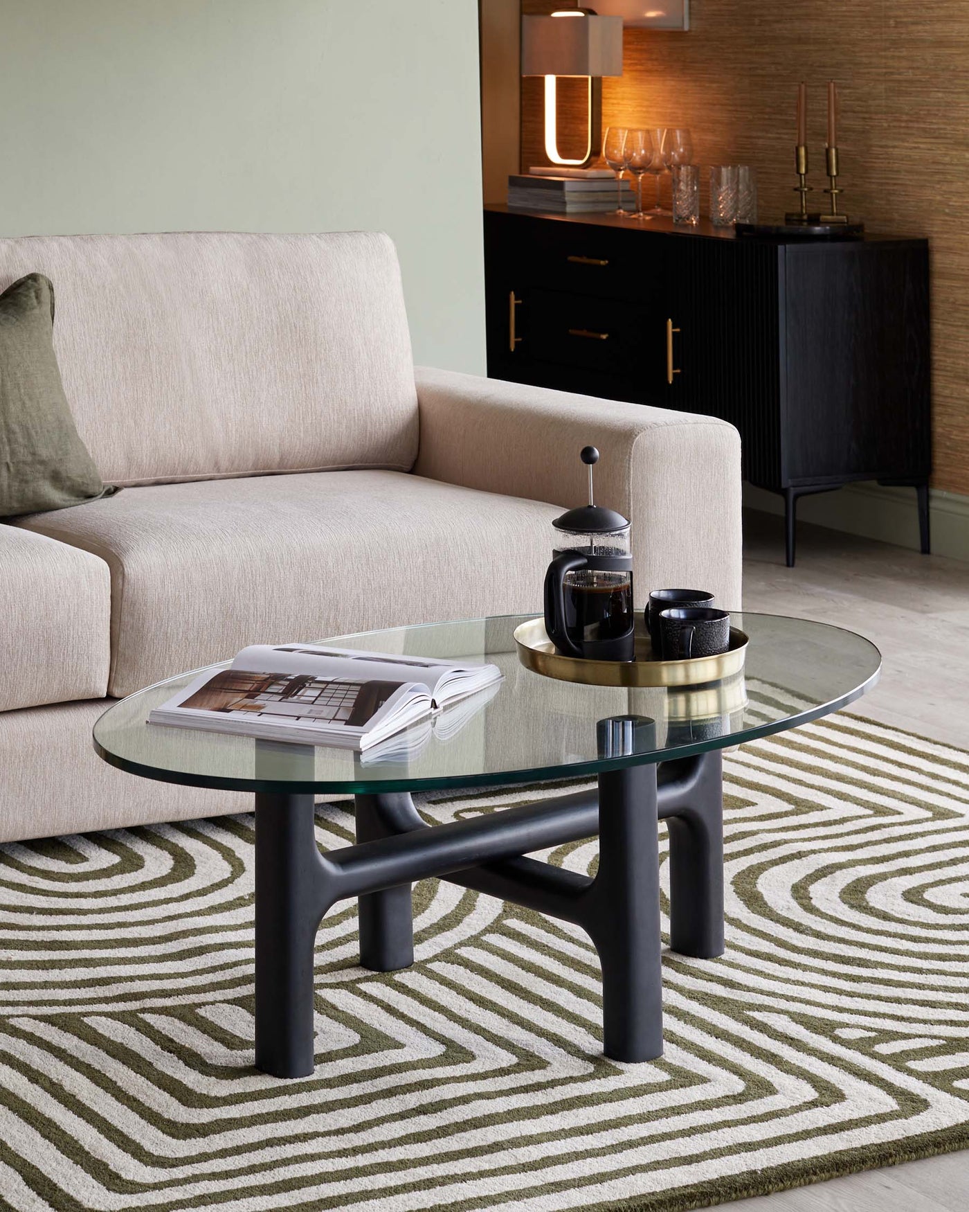 Contemporary living room furniture including a light beige fabric sofa with clean lines and a single green cushion, a round glass coffee table with a unique dark frame, and a black sideboard cabinet with textured doors and brass handles in the background. The ensemble is placed on a patterned beige and green area rug.