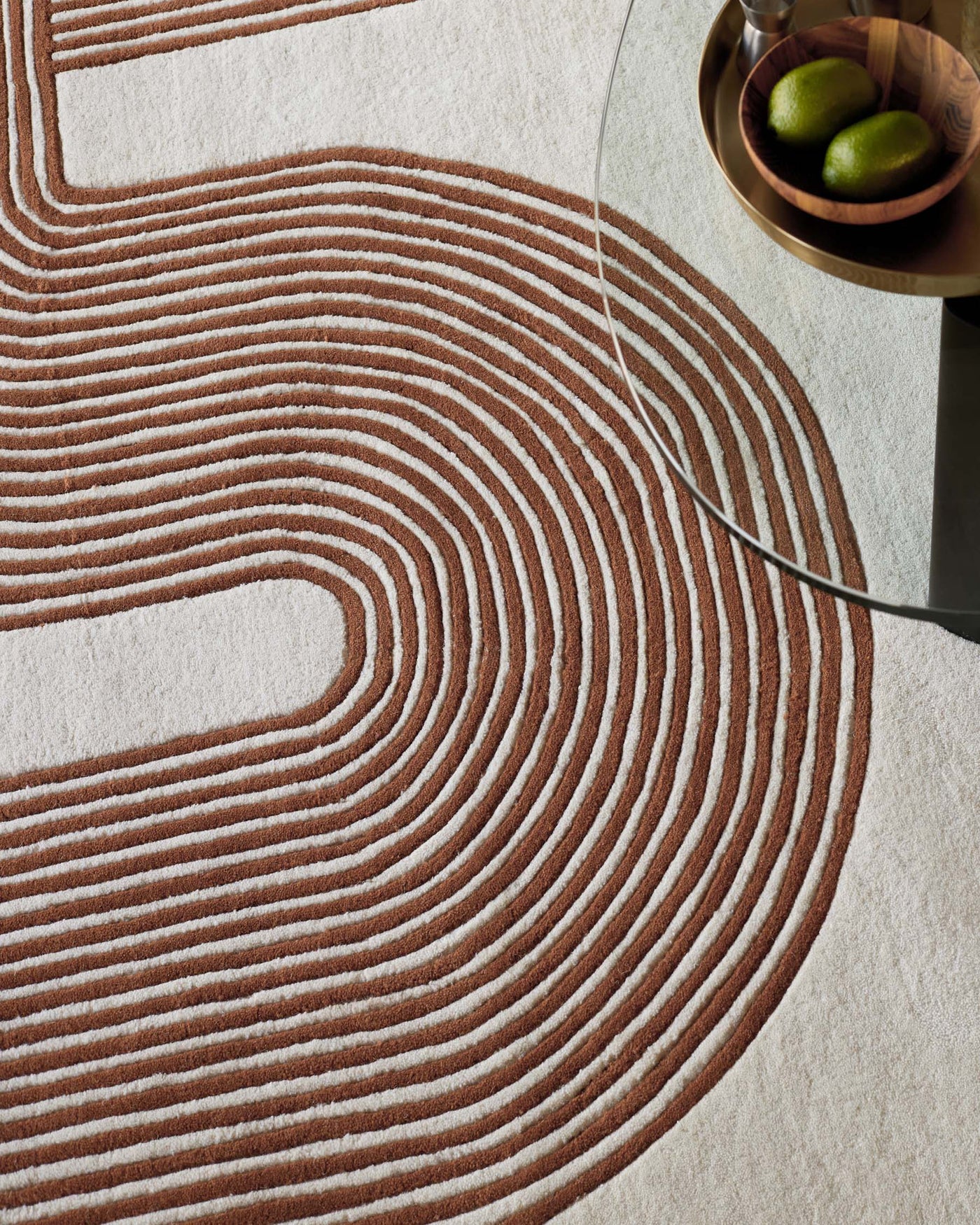 Abstract design area rug with concentric circle pattern in brown and beige, partially under a minimalist glass side table with a round, wooden bowl containing two green limes on top.