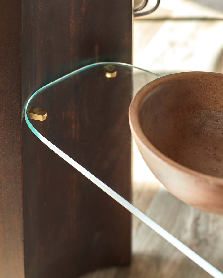 Close-up of a dark wood side table with a glass shelf held up by brass fixtures, with a wooden bowl on top.