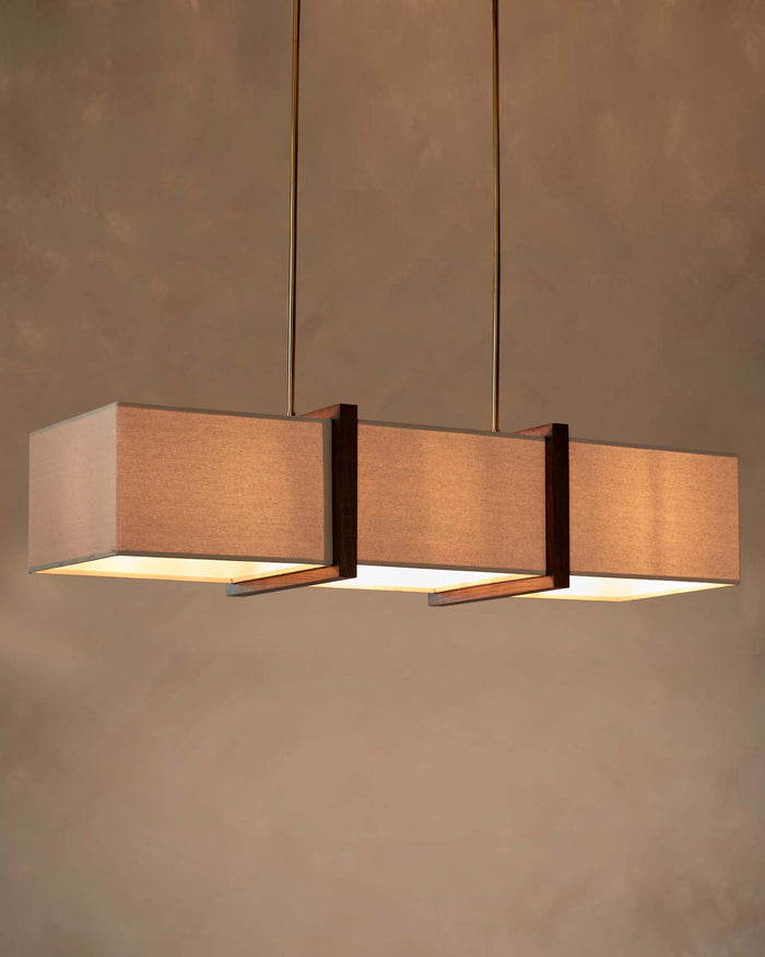 A striking, contemporary linear light fixture, featuring three rectangular shades and a dark wood frame suspended from a gold rod.