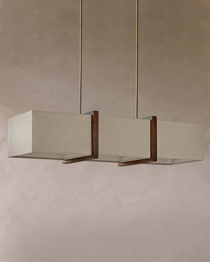 A modern, linear pendant light fixture with three beige fabric shades and warm wood accents, suspended by thin brass rods.