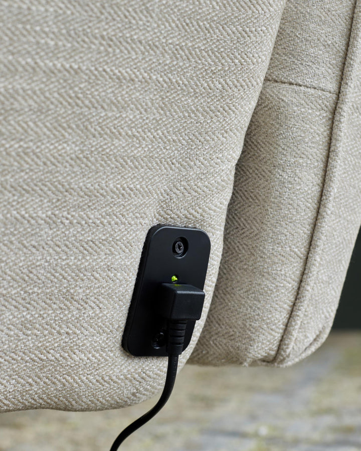 Close-up of a beige upholstered chair with a black power cable and adaptor plugged into the side, illuminated with a green light.