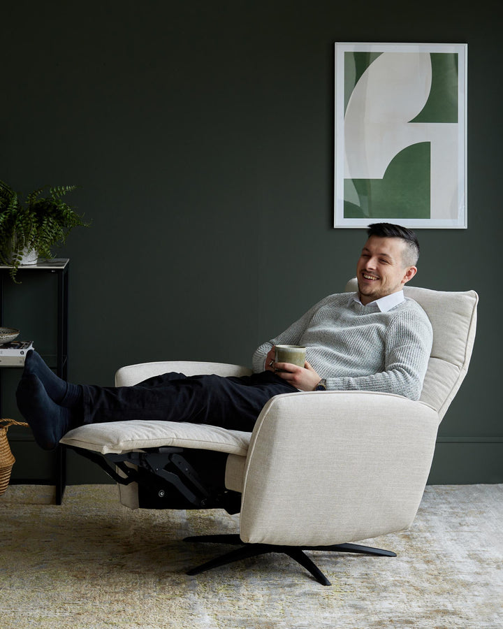 Relaxing recliner chair. Light upholstery, dark legs, with a man sat in it enjoying a cup of tea.