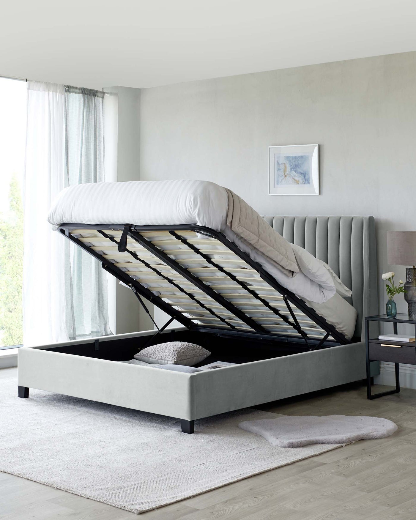 A modern, grey upholstered storage bed with a lifted mattress base revealing a spacious storage area. The bed features a vertical channel-tufted headboard and a sleek, flat upholstered frame resting on short black legs. It is complemented by a grey textured rug and a small, stylish bedside table with a dark finish and metallic frame.