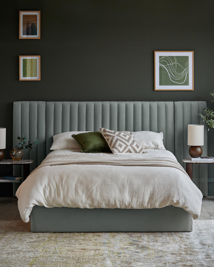 A styled bedroom featuring a bed with a padded headboard, side tables and framed artwork on a dark green wall.