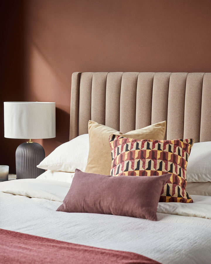 A stylish bed with a brown headboard, adorned with cushions in complementary colours and a bedside lamp.