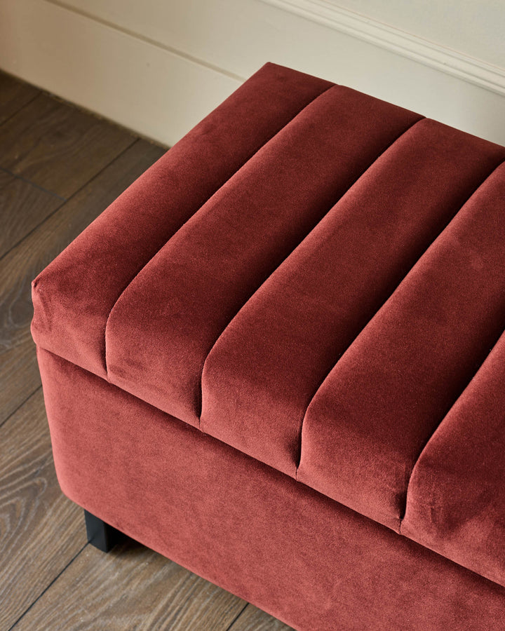 Amalfi velvet ottoman bench in toffee red, shown from angled view displaying channeled back cushions and dark legs on wood flooring.