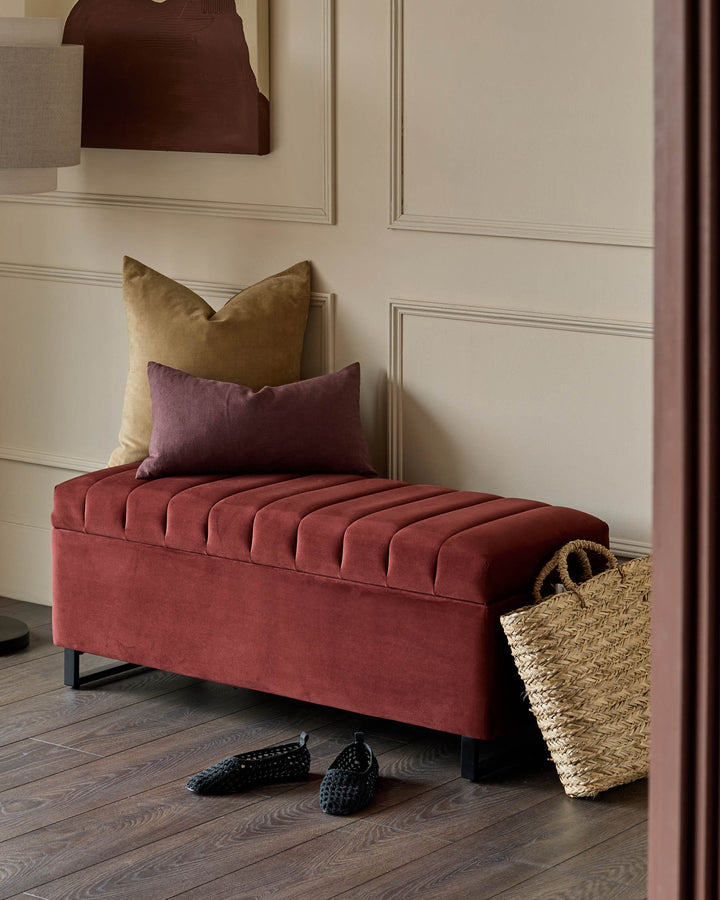 Amalfi velvet ottoman bench in toffee red with pleated top, styled with gold and purple pillows in a modern room setting.