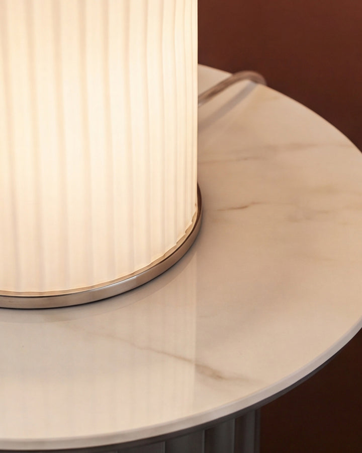 White pleated cylindrical lampshade with brass trim detail, shown at an angle on a round marble table base.