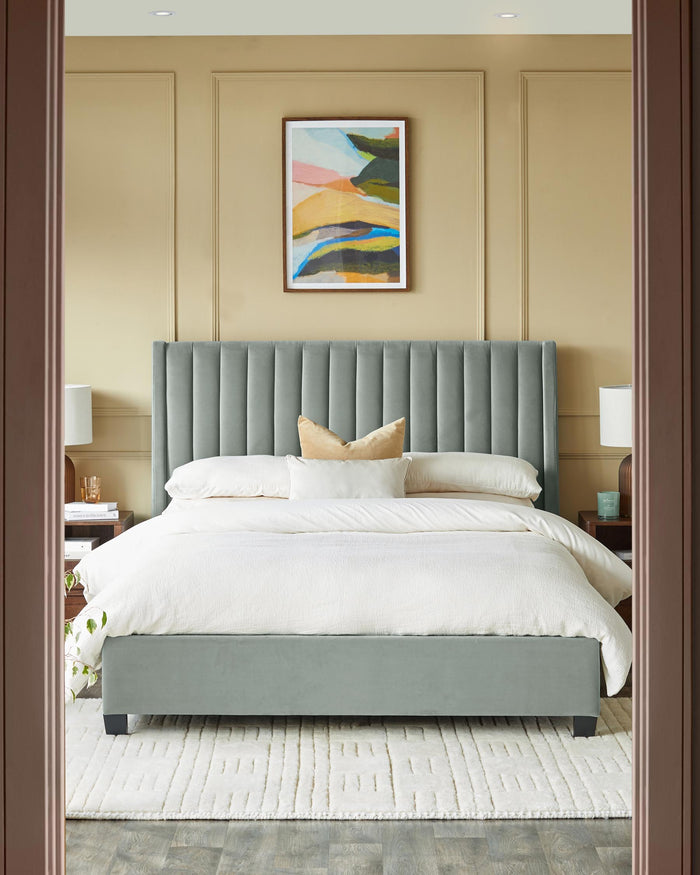 Bedroom with grey velvet bed, white rug and framed artwork above the bed. Matching bedside tables and lamps on either side.