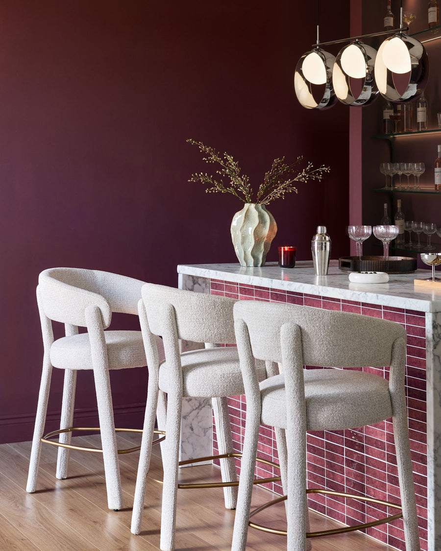 Anya bar stool in natural fabric with curved backrest and brass footrest, shown in burgundy bar setting.