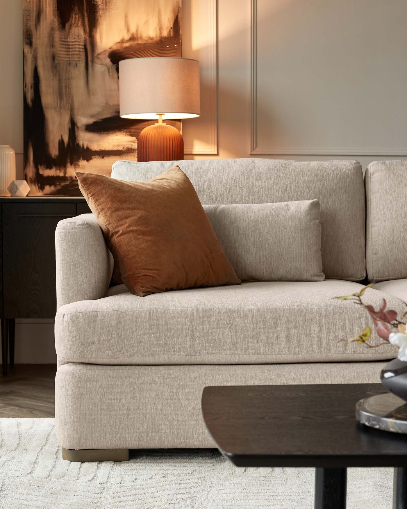 A cosy beige contemporary sofa with a textured fabric is adorned with a plush cinnamon brown throw pillow. Complementing the seating arrangement is a dark wooden coffee table with a rounded triangular top, set on a textured off-white area rug. A modern table lamp with a warm-toned ceramic base and a cream lampshade casts a soft glow, placed on a sleek black sideboard in the background featuring subtle panel detailing. An abstract wall art with earthy tones adds a sophisticated touch to the decor.