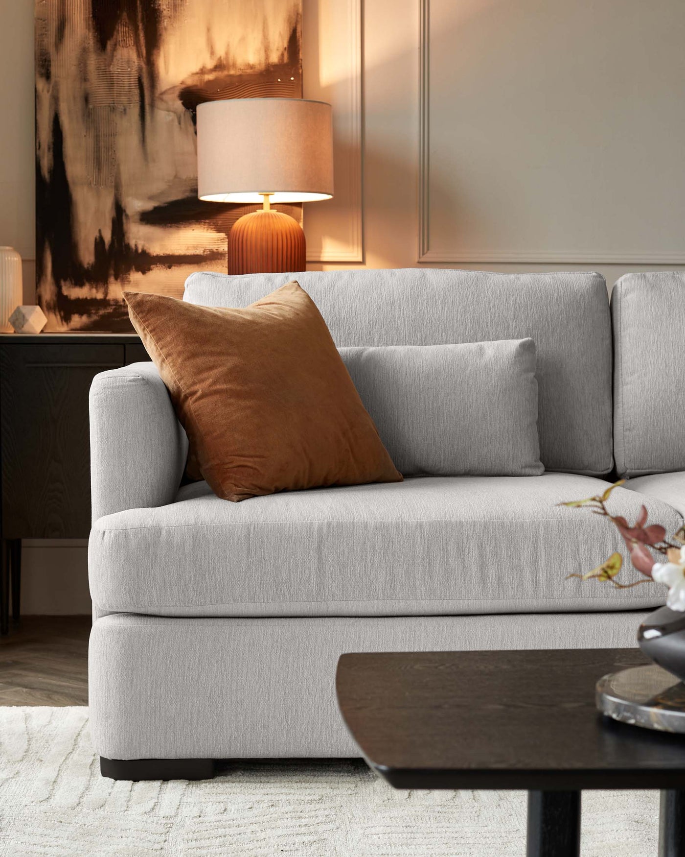 A contemporary living room setting featuring a light grey fabric sofa with clean lines and plush cushions. The sofa is adorned with one terracotta-coloured throw pillow. In the foreground, a dark wooden coffee table with a rounded triangular shape provides contrast. Behind the sofa, a stylish table lamp with a ribbed ceramic base and a beige shade creates a warm ambiance. The furniture arrangement is placed on a textured off-white area rug, giving the space a cosy and inviting feel.