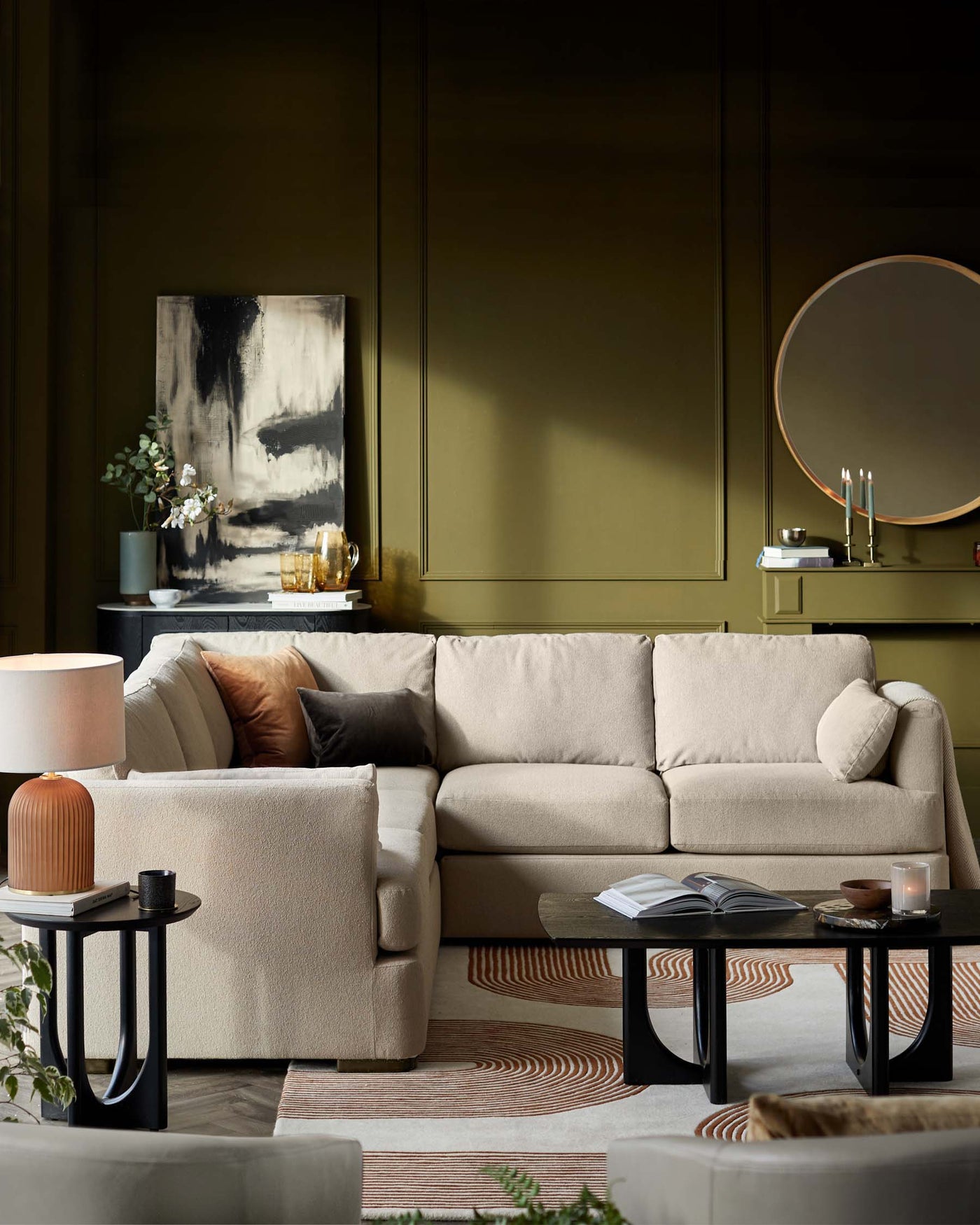 Elegant living room furniture set including a neutral-toned modular sectional sofa with plush cushions, a pair of modern round black side tables, and an abstract-patterned area rug. Accessories include a ceramic table lamp, decorative pillows, and a simple artwork above a sleek console table accompanied by a round mirror.