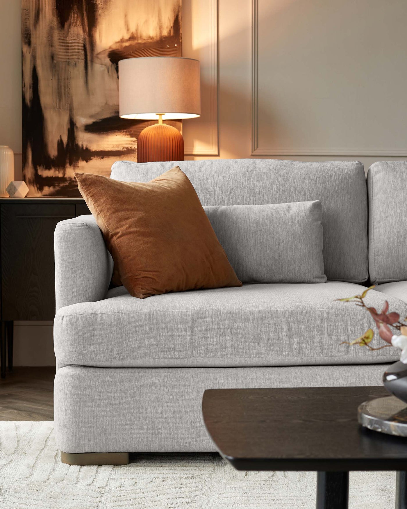 Contemporary light grey fabric sofa with plush cushions and a single brown decorative pillow, paired with a dark wooden coffee table in an understated modern design, situated on a textured off-white area rug. A black wooden sideboard and a stylish table lamp with an orange base and a beige shade enhance the warm and inviting ambiance of the living space.