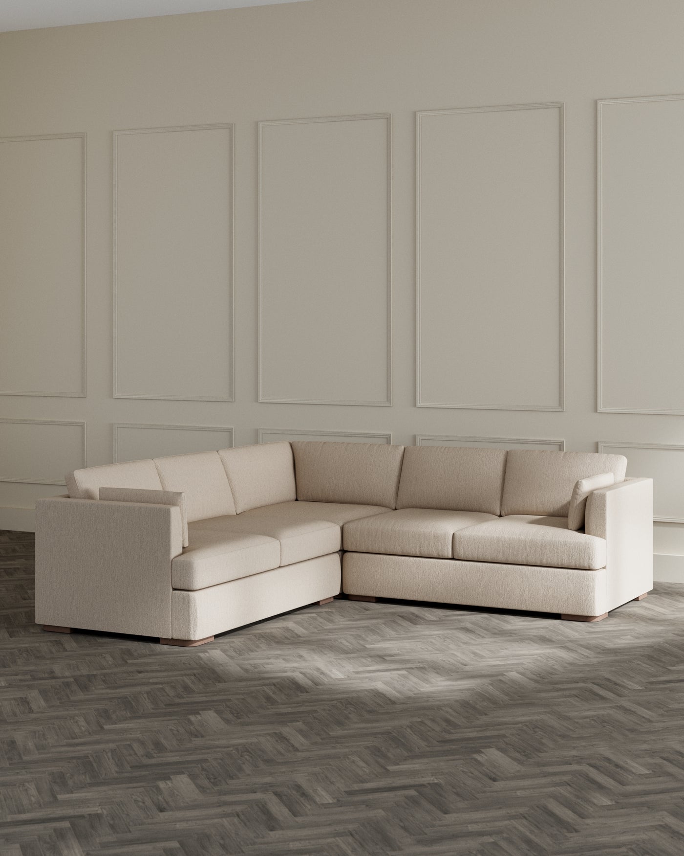 Modern beige L-shaped sectional sofa with clean lines and plush cushions, set against a panelled beige wall on a dark herringbone-patterned floor.