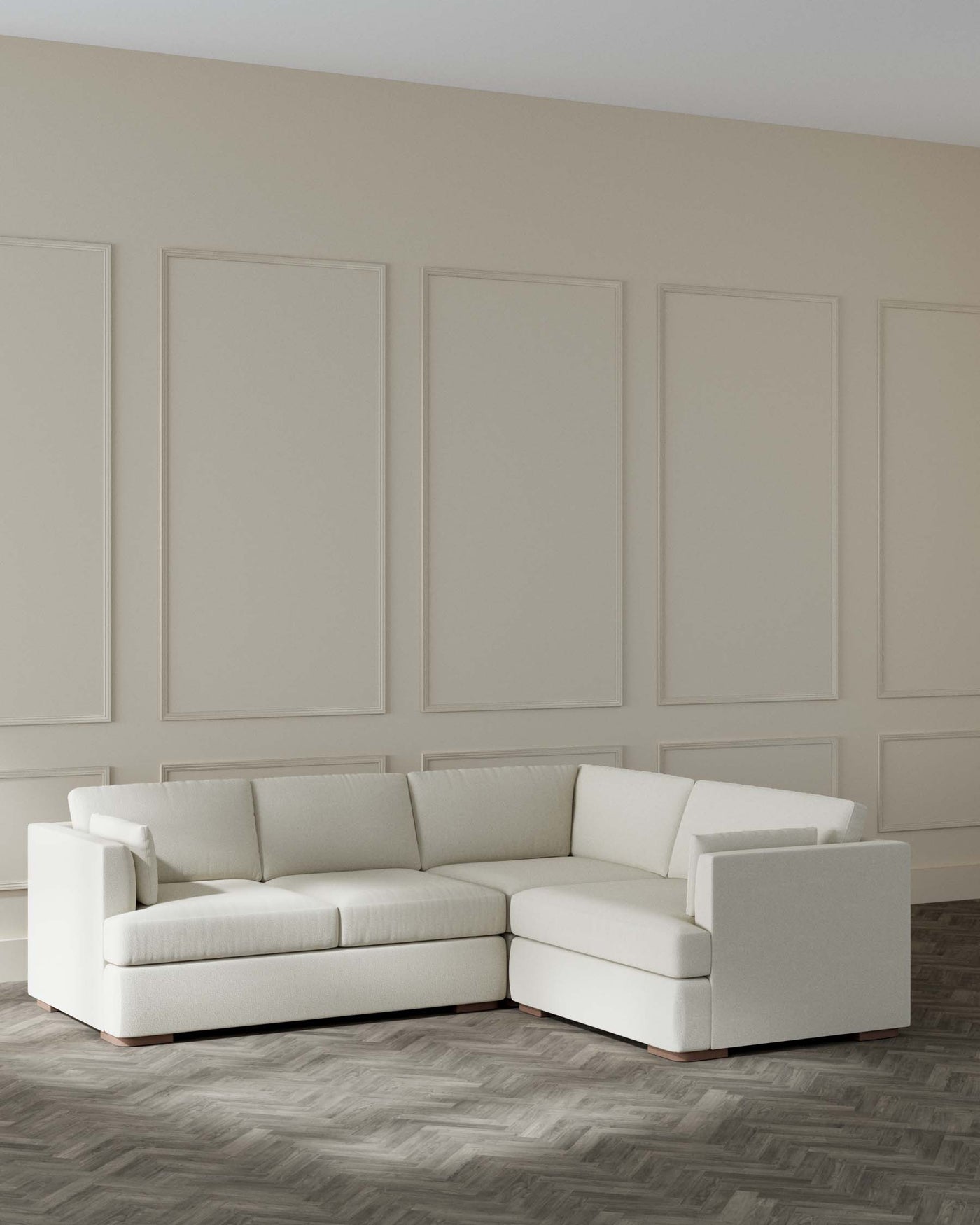 L-shaped modern sectional sofa with a clean, minimalist design, upholstered in light beige fabric, featuring plush cushions and a low-rise back, set on a wood herringbone pattern floor against a panelled wall.