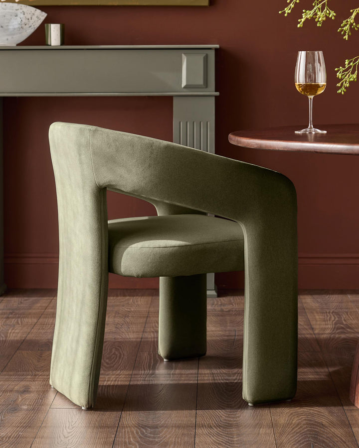 Asher dining chair in olive green fabric, angled side view showing curved armrests and cylindrical base in modern interior setting.