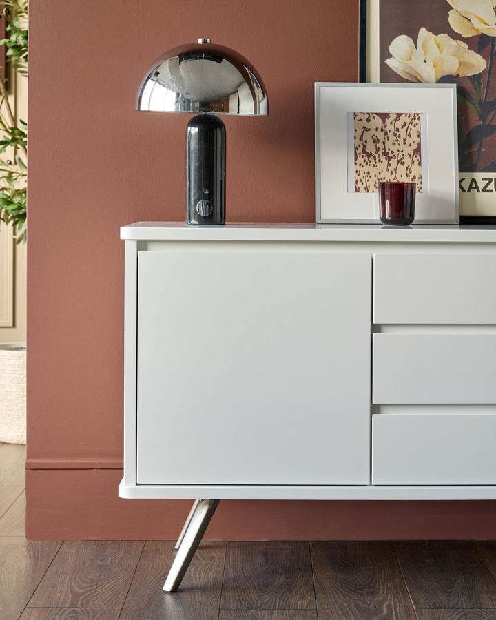 White gloss compact sideboard with angled chrome legs, featuring closed drawers with black handles, displayed against terracotta wall.