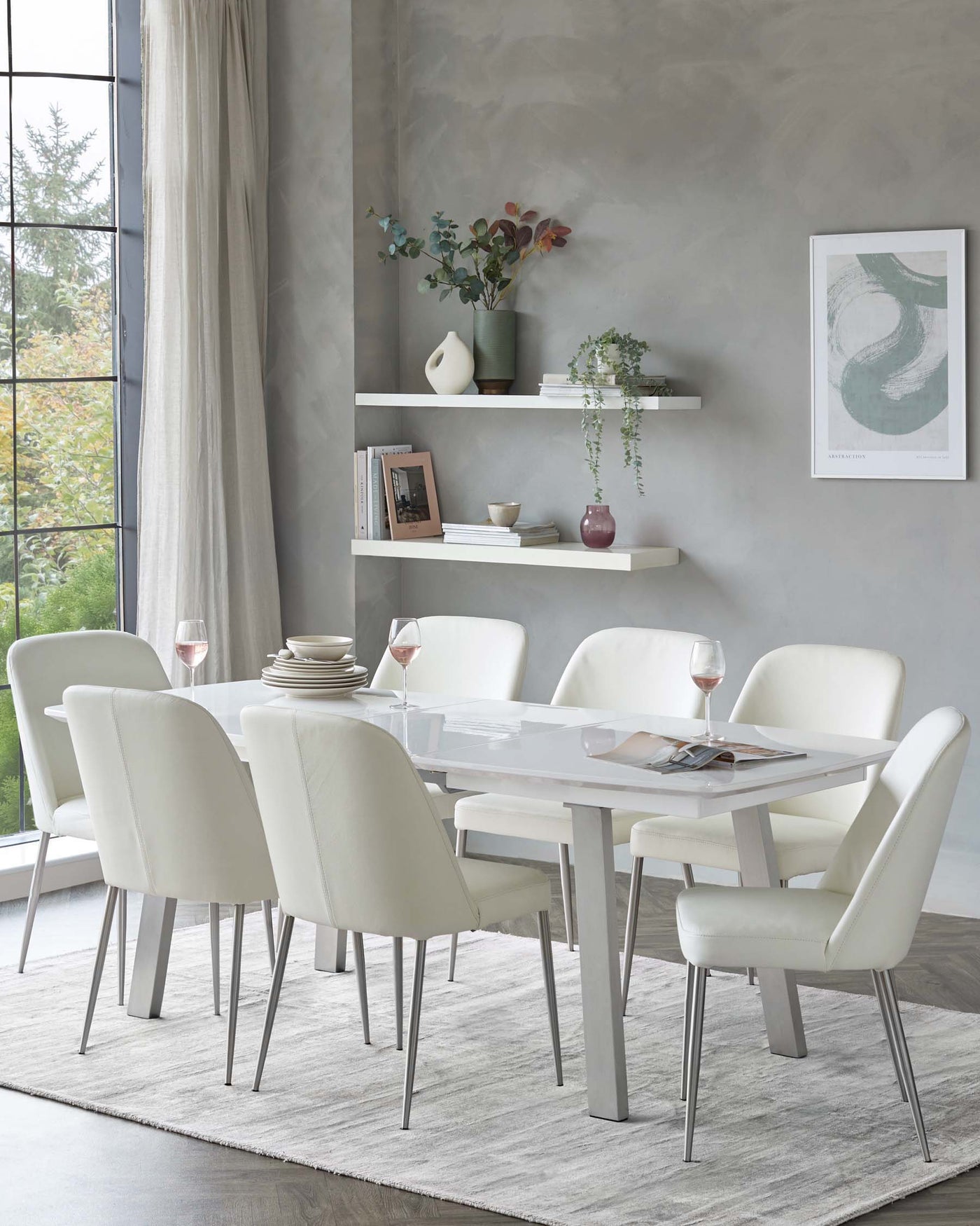 A modern dining room set featuring a rectangular white high-gloss table with silver metal legs, surrounded by six cream upholstered dining chairs with sleek metal legs. The table is partially set with plates and wine glasses. A light grey area rug underlines the set, and a couple of white floating shelves holding decorative items are mounted on the wall behind.