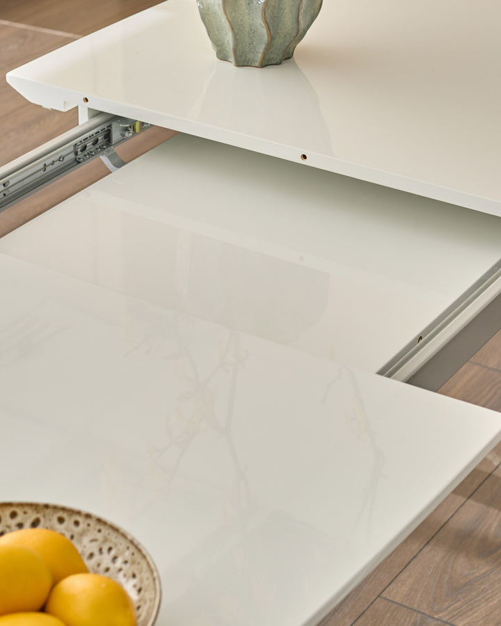 White extending dining table with open drawer, showing metal slide mechanism and glossy finish, styled with cactus and lemons.
