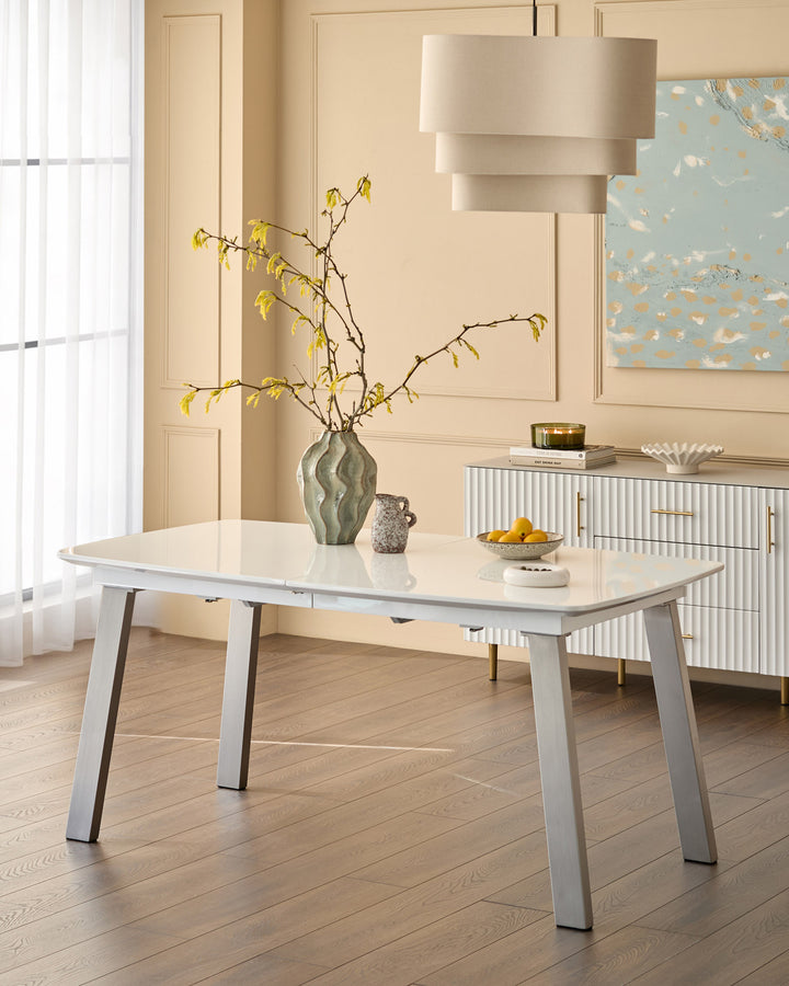 White extending dining table with grey metal legs, displayed in a light modern dining room with beige walls and wooden flooring.