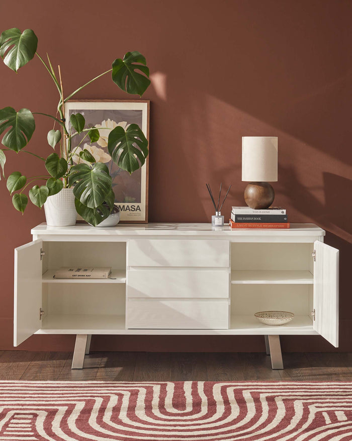 A white sideboard with drawers and cupboards sits against a warm brown wall, accessorised with a lamp, books, and a plant.