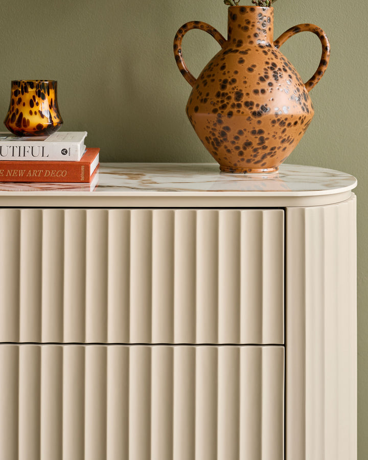 Austin chest of drawers in natural ceramic with vertical fluted detailing and rounded marble top, styled with decorative objects.