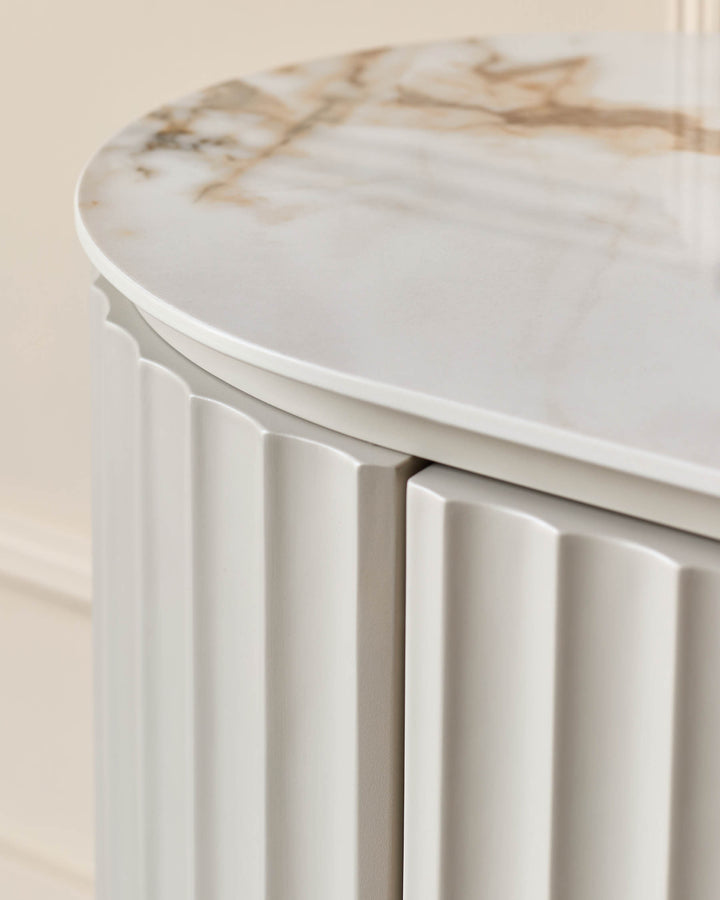 A modern bedside table in cream with a marble top and fluted panels. It features a round top and sleek design.