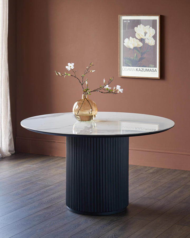 A modern round marble dining table with a ribbed, dark blue pedestal base. A vase of flowers sits on top.