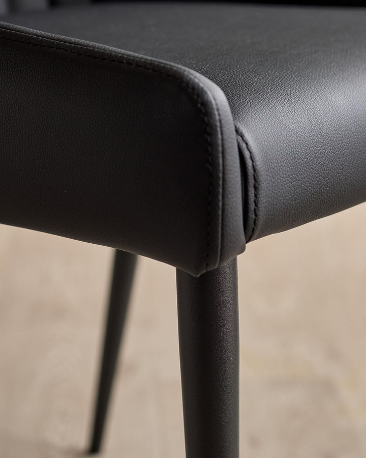 Close-up of black faux leather dining chair showing textured seat padding and dark metal leg detail.