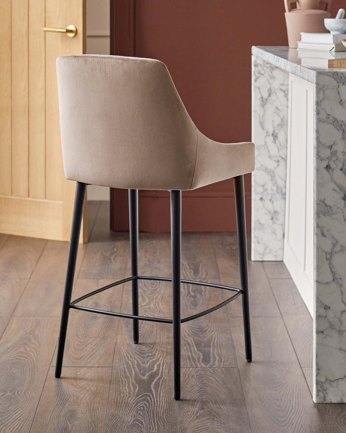 A beige bar stool with black metal legs sits on a wooden floor in front of a marble topped kitchen counter.