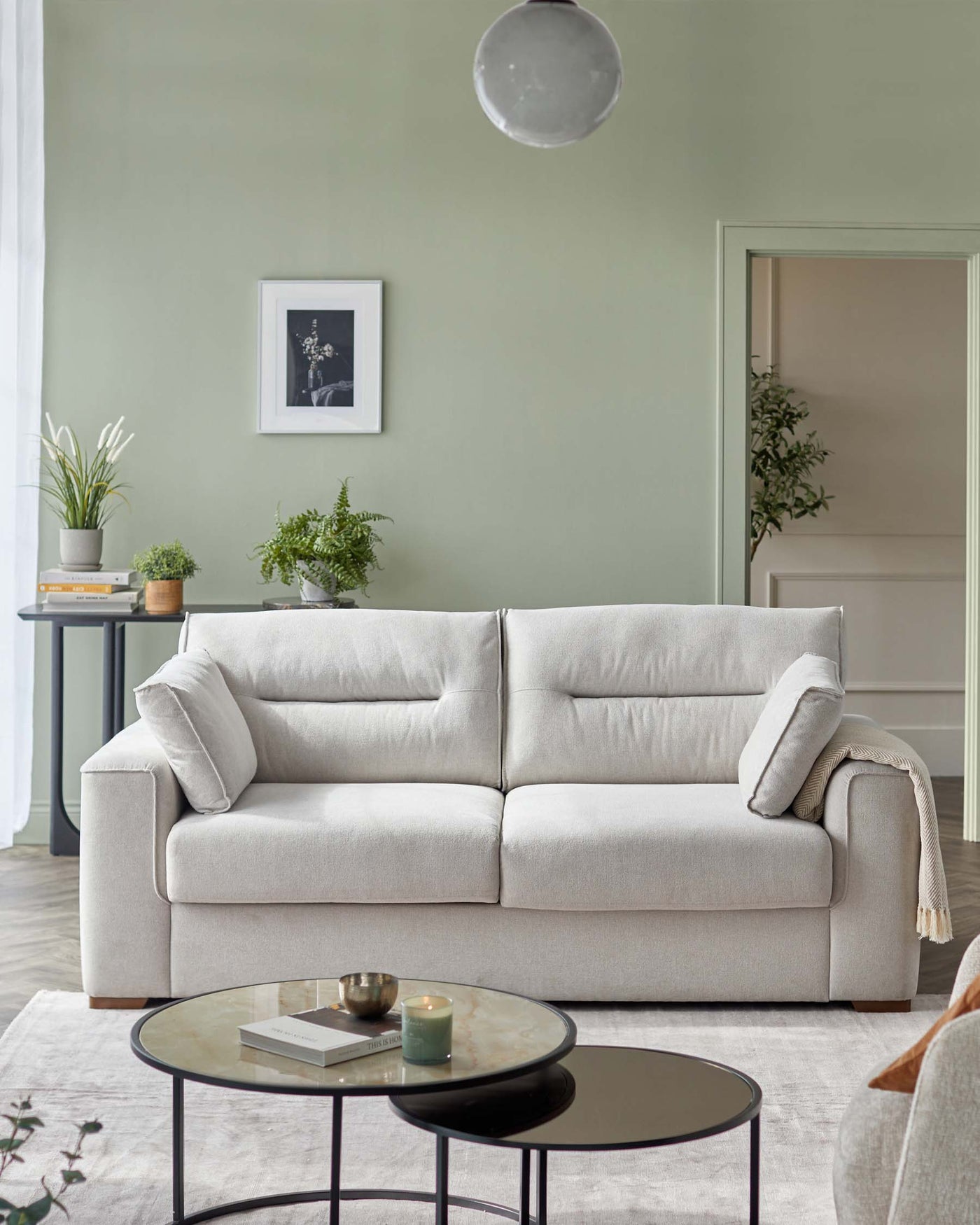 Modern light grey three-seater fabric sofa with plush cushions and simple armrests, paired with a round, nesting-style coffee table set featuring a black metal frame and dark glass top on the larger table and a marble effect top on the smaller table, showcased in a contemporary living room setting with soft lighting and neutral tones.