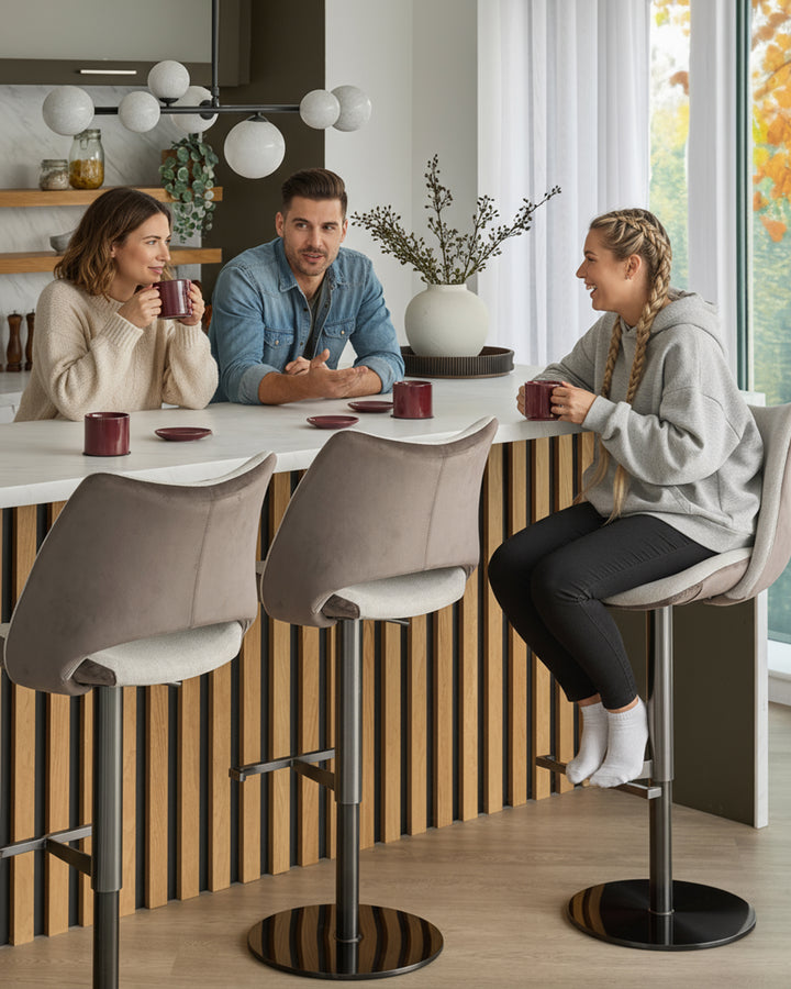 Bronwyn gas lift bar stool in natural and mocha velvet, shown in kitchen setting with three people seated at counter.