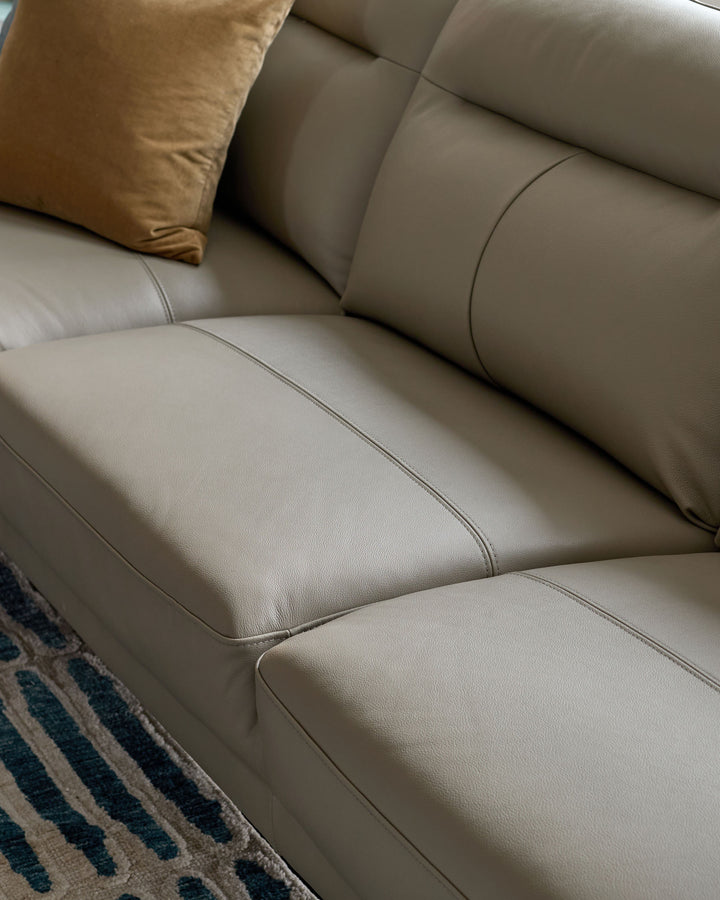 Light grey leather left-hand corner chaise sofa with cushioned seating and bronze pillow accent, angled view.