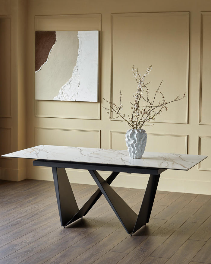 A modern marble-topped dining table with striking black, geometric legs, and a white vase with floral arrangement.
